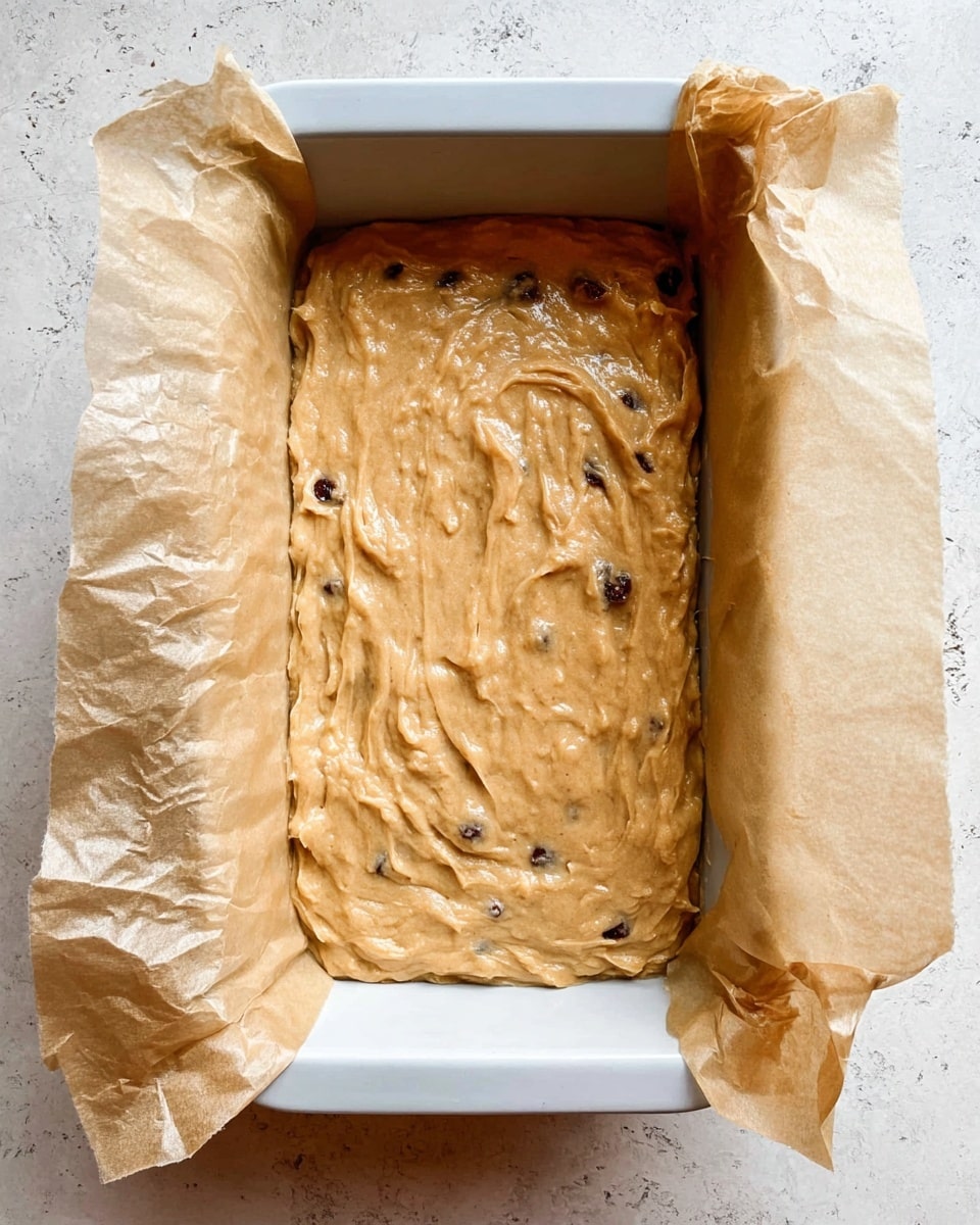 The image shows a rectangular white baking pan lined with crinkled light brown parchment paper that extends above the pan’s edges. Inside, there is one uneven layer of light golden brown thick batter with a few scattered dark spots, which look like raisins or chocolate chips, spread roughly from edge to edge. The baking pan is set on a white marbled surface with slight beige veining. The texture of the batter is slightly lumpy and smooth in some places, with gentle waves and streaks from spreading. Photo taken with an iphone --ar 4:5 --v 7