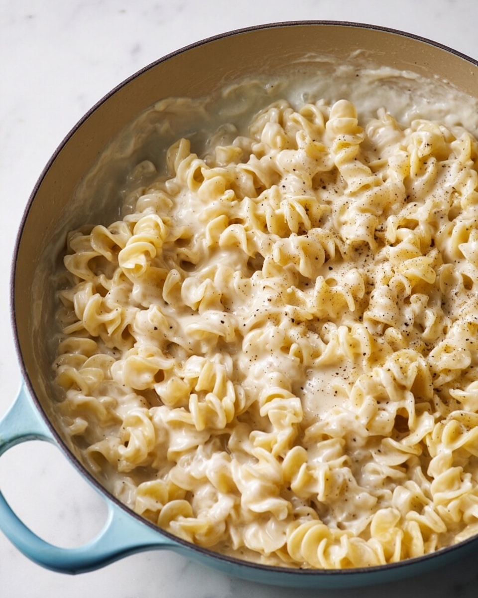 A close-up view of a light blue pan filled with creamy, curly pasta coated in a thick white sauce with specks of black pepper scattered throughout. The pasta is placed evenly across the pan, sitting on a white marbled surface. The sauce clings to the pasta, giving it a smooth and rich texture while the pan's handle remains visible on the left side. Photo taken with an iphone --ar 4:5 --v 7