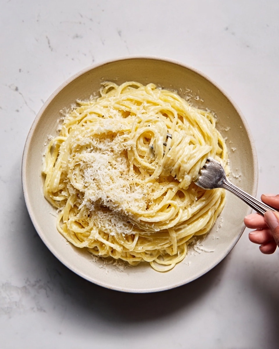A mound of creamy spaghetti noodles piled high in a white bowl sits on a white marbled surface. The spaghetti looks smooth and coated evenly with a pale, white sauce, speckled lightly with black pepper. Two silver forks are stuck into the pasta, positioned at slightly different angles, with sauce clinging to them. The noodles are thick and glossy, winding around each other in soft spirals. photo taken with an iphone --ar 4:5 --v 7
