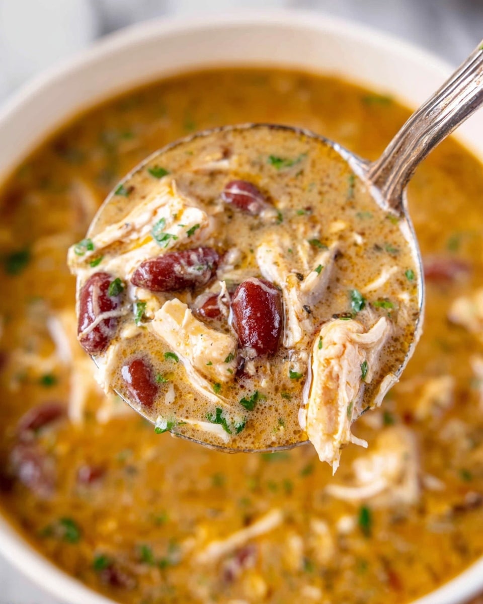 A close-up view of a ladle filled with creamy soup showing several layers including shredded light beige chicken pieces, dark red kidney beans, and small bits of green herbs sprinkled on top. The soup base is thick and light brown with a smooth, slightly oily texture. The ladle is shiny silver and is held above a blurred white bowl filled with the same soup, resting on a white marbled surface. photo taken with an iphone --ar 4:5 --v 7