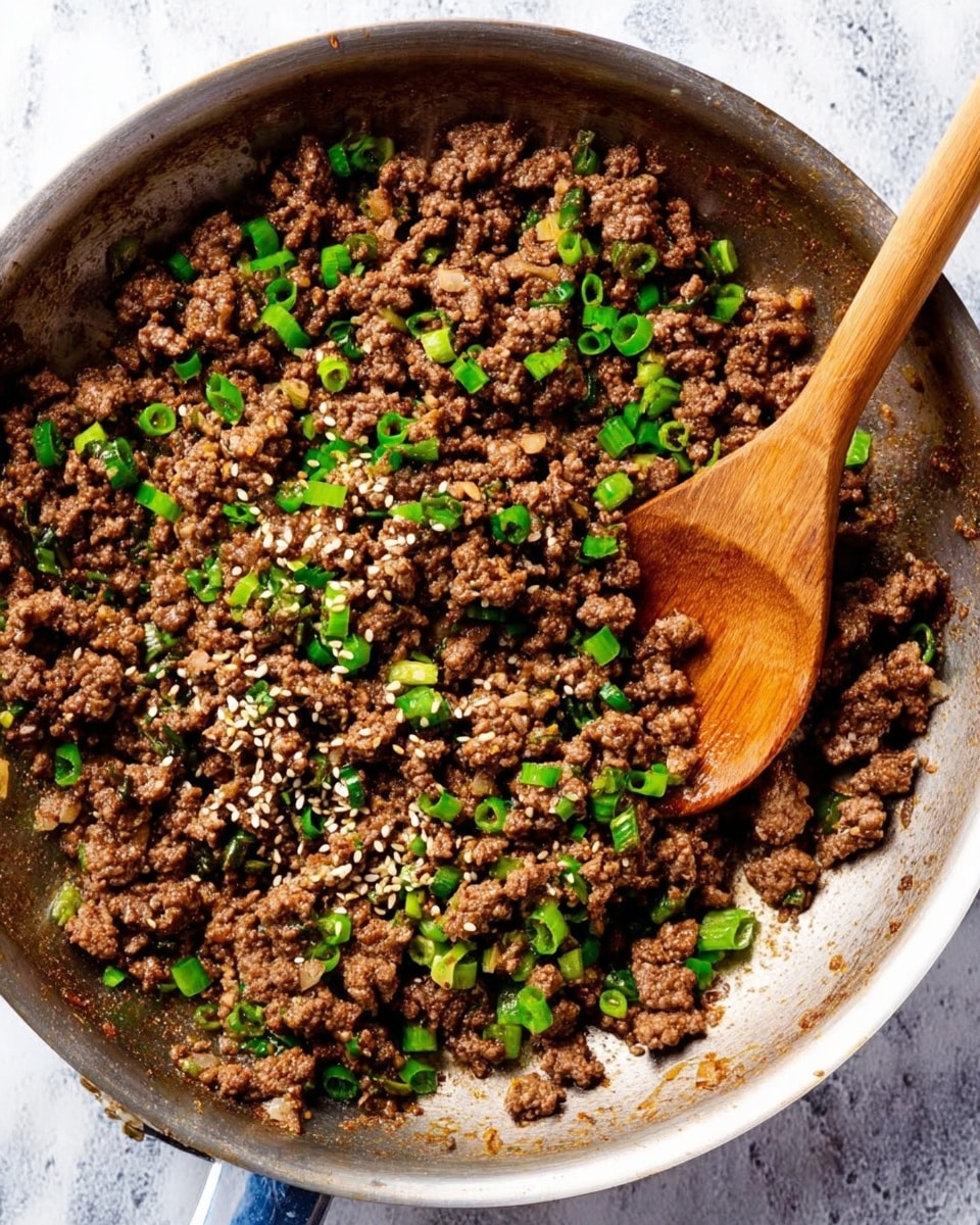 A close-up top view of a silver pan filled with cooked ground beef, showing a crumbly texture with small pieces of browned meat mixed with green chopped scallions and light sesame seeds scattered on top. A wooden spoon rests inside the pan on the right side. The pan sits on a white marbled surface. photo taken with an iphone --ar 4:5 --v 7