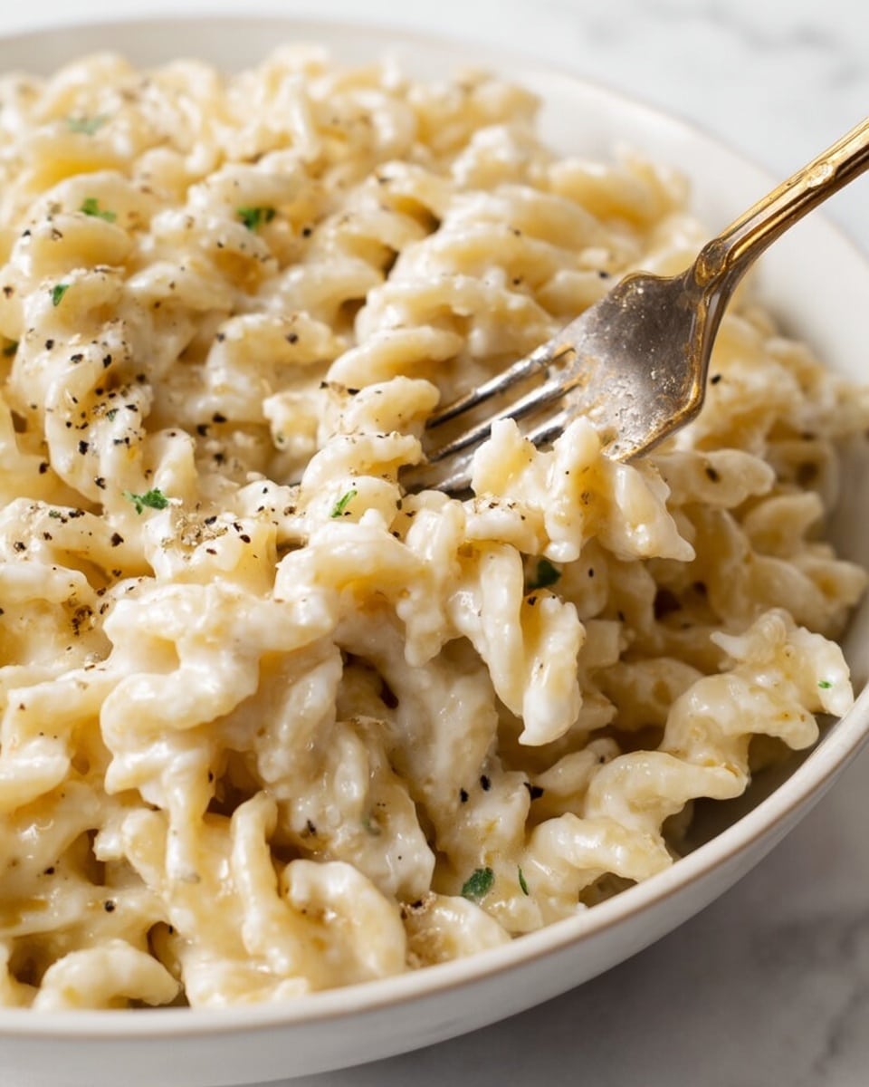 The image shows a close-up of creamy pasta in a white bowl, with a fork partially inserted into the noodles. The pasta has short, twisted shapes with a smooth white sauce coating each piece evenly. Small green herb bits and black pepper flakes are scattered across the pasta, adding specks of color and texture. The background is a white marbled surface. Photo taken with an iphone --ar 4:5 --v 7