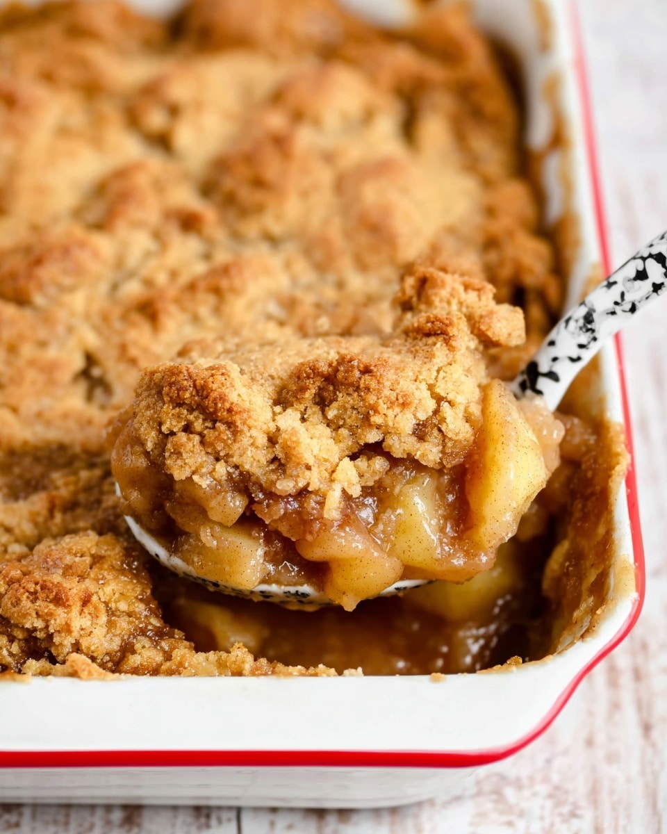 The image shows a close-up of a warm apple cobbler inside a white baking dish with a red rim. The cobbler has two clear layers: the bottom layer is made up of soft, juicy, golden-brown cooked apples with a smooth texture, while the top layer is a crumbly, golden-brown crust with uneven, cracked pieces that look slightly crispy. A white spoon with black marbled patterns is scooping a chunk of the cobbler out, revealing the gooey filling underneath the crust. The dish sits on a white marbled surface, and the focus is on the spoonful in the foreground. Photo taken with an iphone --ar 4:5 --v 7