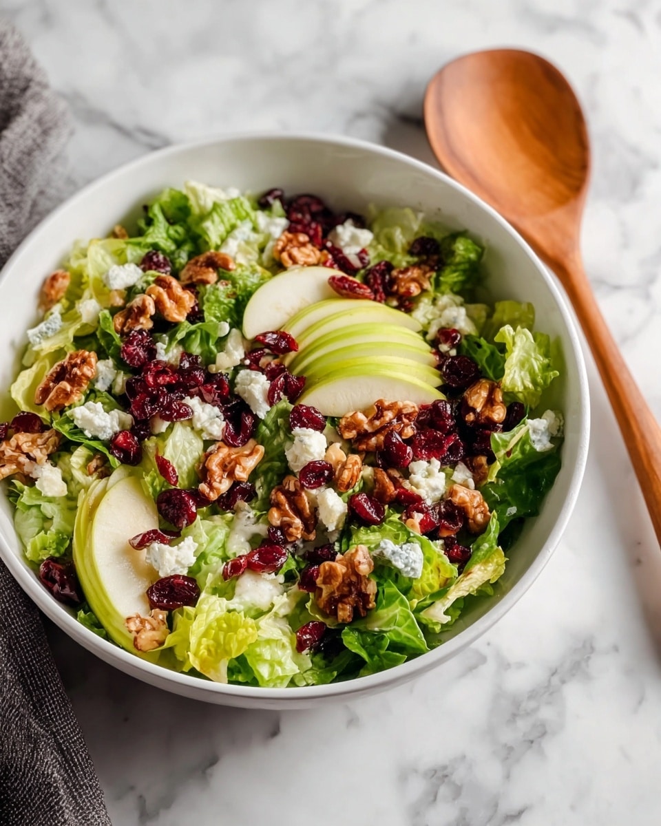 A white bowl filled with a fresh salad on a white marbled surface. The salad has several layers starting with bright green lettuce leaves at the base, topped with light green apple slices, deep red dried cranberries, and many brown walnut pieces. Scattered small, soft dollops of white cheese are sprinkled throughout the salad, adding texture. A wooden spoon lies beside the bowl. Photo taken with an iphone --ar 4:5 --v 7