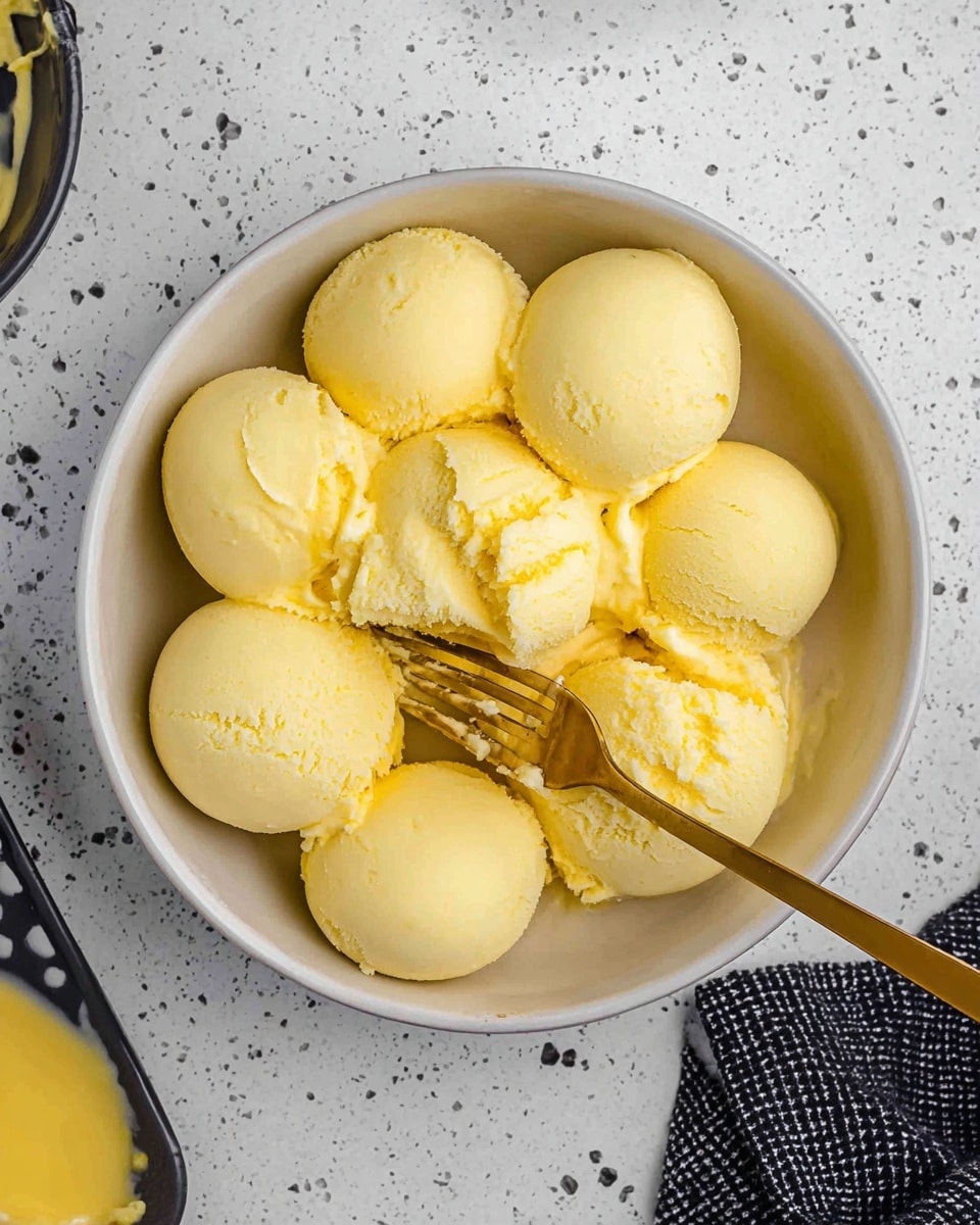 The image shows a white bowl filled with about a dozen yellow, smooth, rounded scoops that appear creamy and soft, some broken into chunks. A golden fork is pressing into the scoops in the center of the bowl, breaking some apart and creating a rough texture in the middle. The bowl sits on a white marbled surface with small black specks and dots all over. Part of another dish with a yellow sauce can be seen at the bottom left, and a black and white checkered cloth is partly visible at the bottom right. photo taken with an iphone --ar 4:5 --v 7