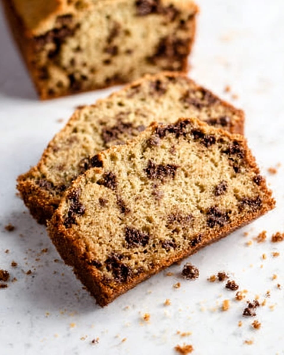 The image shows two slices of banana bread with chocolate chips on a white marbled surface. The bread has a golden brown crust and a moist texture with visible chocolate pieces throughout. One slice rests partly on the other, with crumbs scattered around them, adding a homemade feel. The color of the bread is light brown with darker spots from the chocolate chips. In the background, part of the remaining loaf is out of focus. The lighting gives the bread a warm and inviting look. Photo taken with an iphone --ar 4:5 --v 7