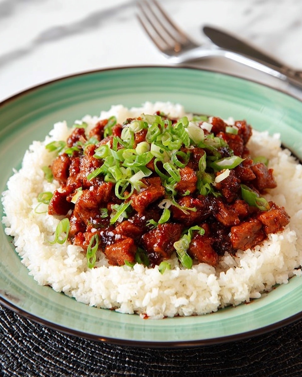 A dish on a white plate with a greenish inner rim, showing three main layers: the bottom layer is soft white rice spread around the plate, forming a ring; the middle layer is reddish-brown saucy pieces of cooked meat piled in the center; the top layer is bright green chopped scallions sprinkled over the meat. The plate rests on a white marbled surface, with a silver fork in the background. photo taken with an iphone --ar 4:5 --v 7