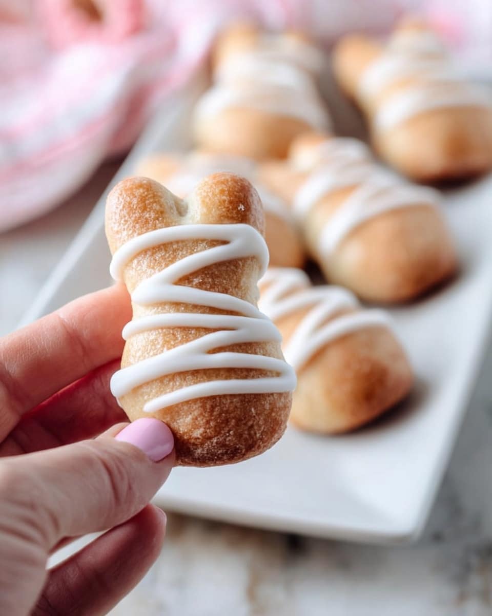 A woman's hand is holding a small bunny-shaped pastry with a golden brown color and a slightly textured surface. The pastry has thick white icing drizzled in diagonal stripes across its body. In the background, several similar pastries lie in a row on a long white plate, all decorated with the same white icing pattern. The setting includes a soft, white marbled surface beneath the plate. photo taken with an iphone --ar 4:5 --v 7