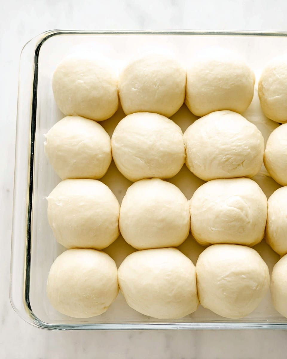 The image shows a clear glass baking dish filled with two neat rows of smooth, round dough balls ready for baking. Each dough ball is pale cream in color with a soft, slightly shiny texture, evenly spaced with small gaps in between. The dough balls have a consistent shape, looking soft and fluffy with subtle creases on their surface. The dish is placed on a white marbled surface, bright and clean, highlighting the light color of the dough. photo taken with an iphone --ar 4:5 --v 7