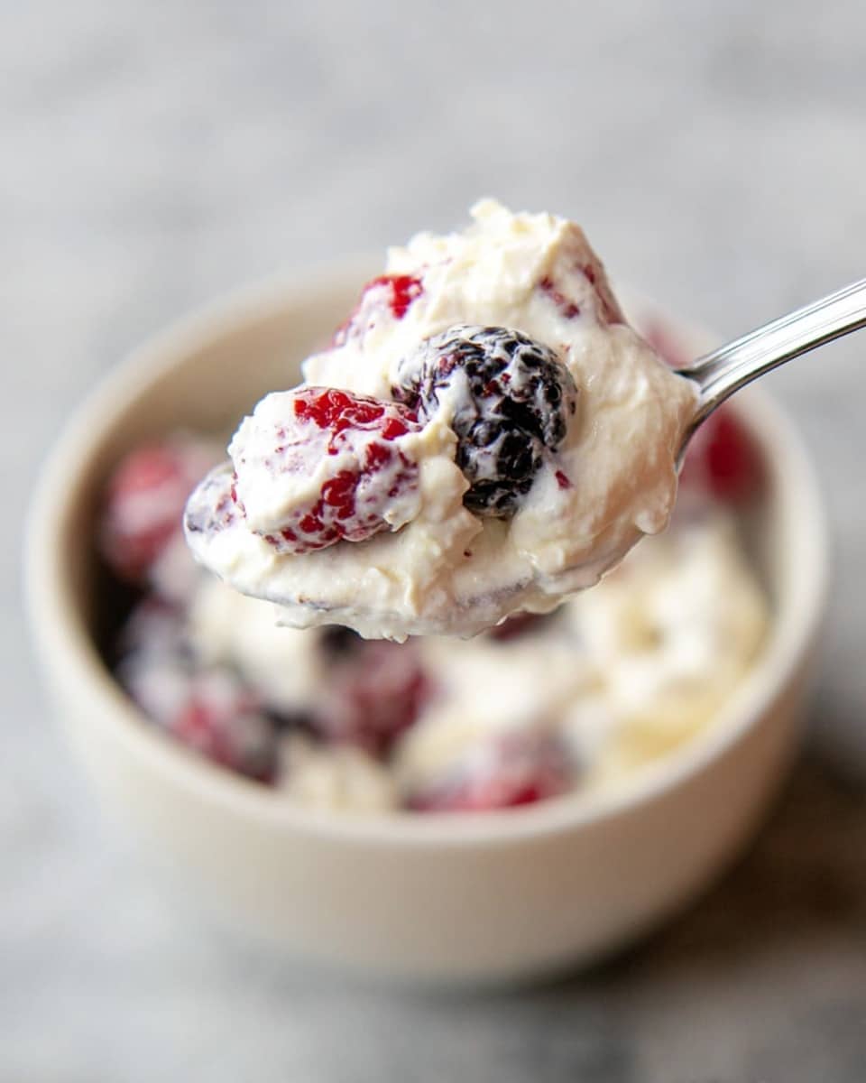A spoon holds a close-up of a creamy white mixture with visible whole blackberries and raspberries, showing a soft, fluffy texture with the berries partially covered in the creamy layer. The spoon is lifted above a white bowl filled with more of the same creamy berry mix, placed on a white marbled surface. The image focuses on the spoonful in the foreground with the bowl blurred in the background. photo taken with an iphone --ar 4:5 --v 7