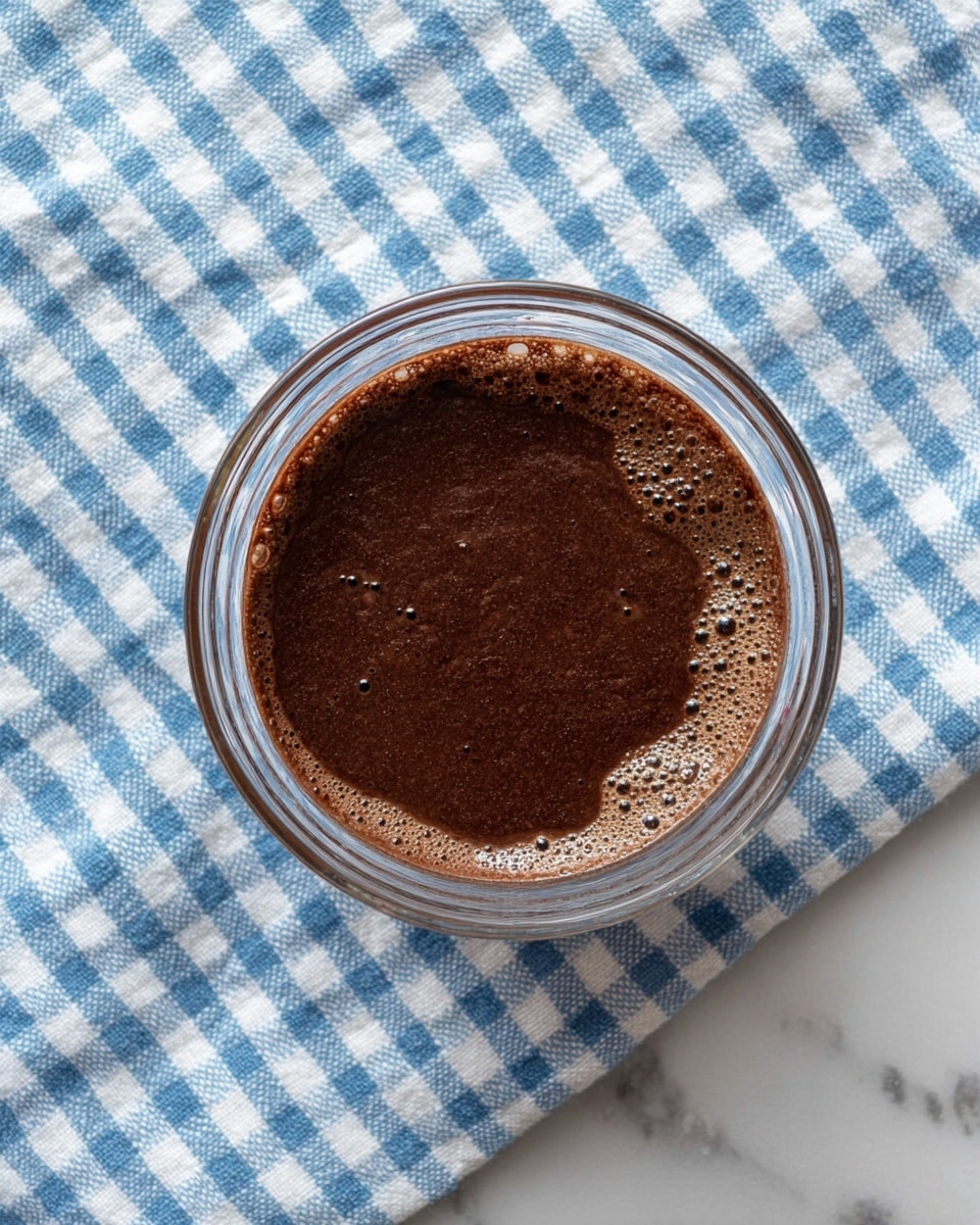 A clear glass bowl containing a single layer of dark brown chocolate liquid mixture with a smooth and slightly bubbly surface, sitting on a white marbled texture with a blue and white checkered cloth underneath. The mixture has some swirls of lighter brown around the edges inside the bowl. photo taken with an iphone --ar 4:5 --v 7