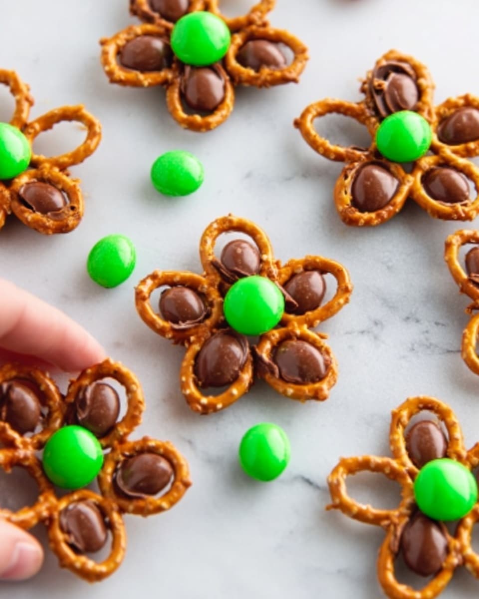 The image shows several flower-shaped treats made of pretzel sticks arranged in a circle on a white marbled surface. Each flower has five pretzel sticks forming the petals, with a round milk chocolate candy placed at the center of the flower. On top of the chocolate candy in the center, there is a single green candy adding a bright pop of color. One flower is being held gently by a woman's hand at the bottom left of the image. Scattered green candies are also visible on the surface around the flowers. photo taken with an iphone --ar 4:5 --v 7