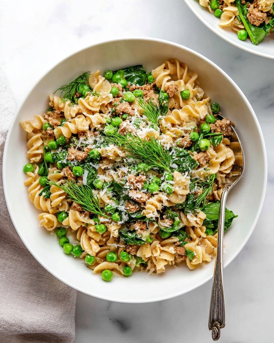 A white bowl filled with curly pasta layered with light brown cooked meat pieces, bright green peas, and fresh green spinach leaves mixed evenly throughout. The top layer has a sprinkle of grated white cheese and scattered small fresh dill leaves. A silver spoon rests inside the bowl on the right side, and the bowl is placed on a white marbled surface with another white bowl partly visible at the top right. Photo taken with an iphone --ar 4:5 --v 7