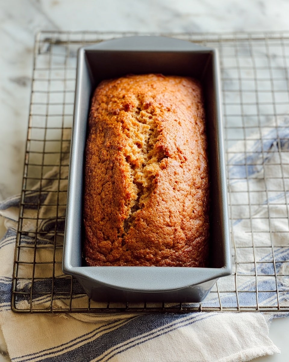 A warm brown loaf of bread with a slightly rough and cracked top surface sits inside a gray metal loaf pan. The bread has a dense texture with a few visible air pockets. The pan rests on a metal cooling rack, which is placed on a white marbled surface covered partially by a blue and beige striped cloth. Photo taken with an iphone --ar 4:5 --v 7