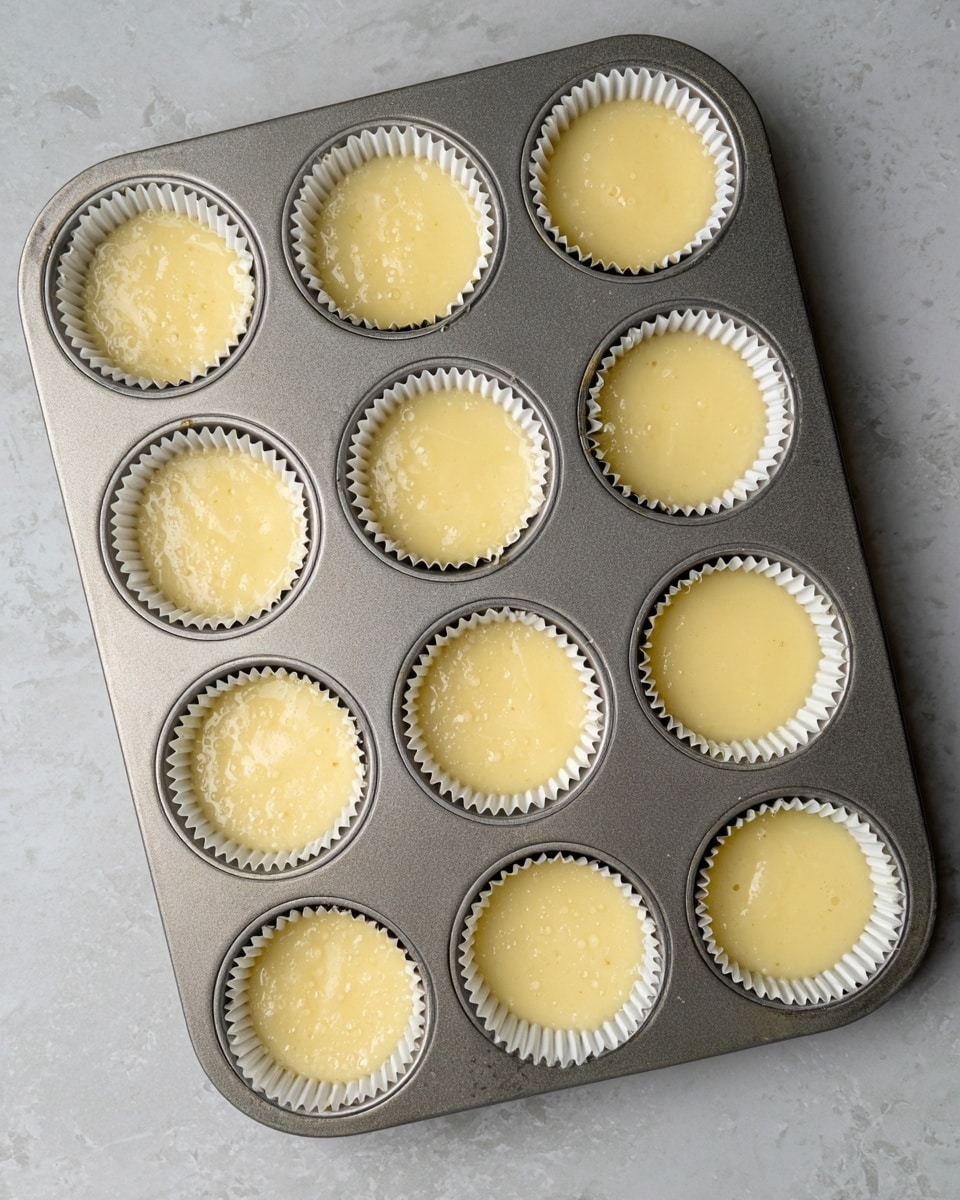 A metallic cupcake tray with twelve round wells, each holding a white paper liner filled with pale yellow cake batter that looks smooth with small bubbles on the surface. The tray rests on a white marbled textured surface, and the batter in the liners reaches nearly to the top edges. The scene is bright with soft lighting, showing a clean kitchen setting. photo taken with an iphone --ar 4:5 --v 7