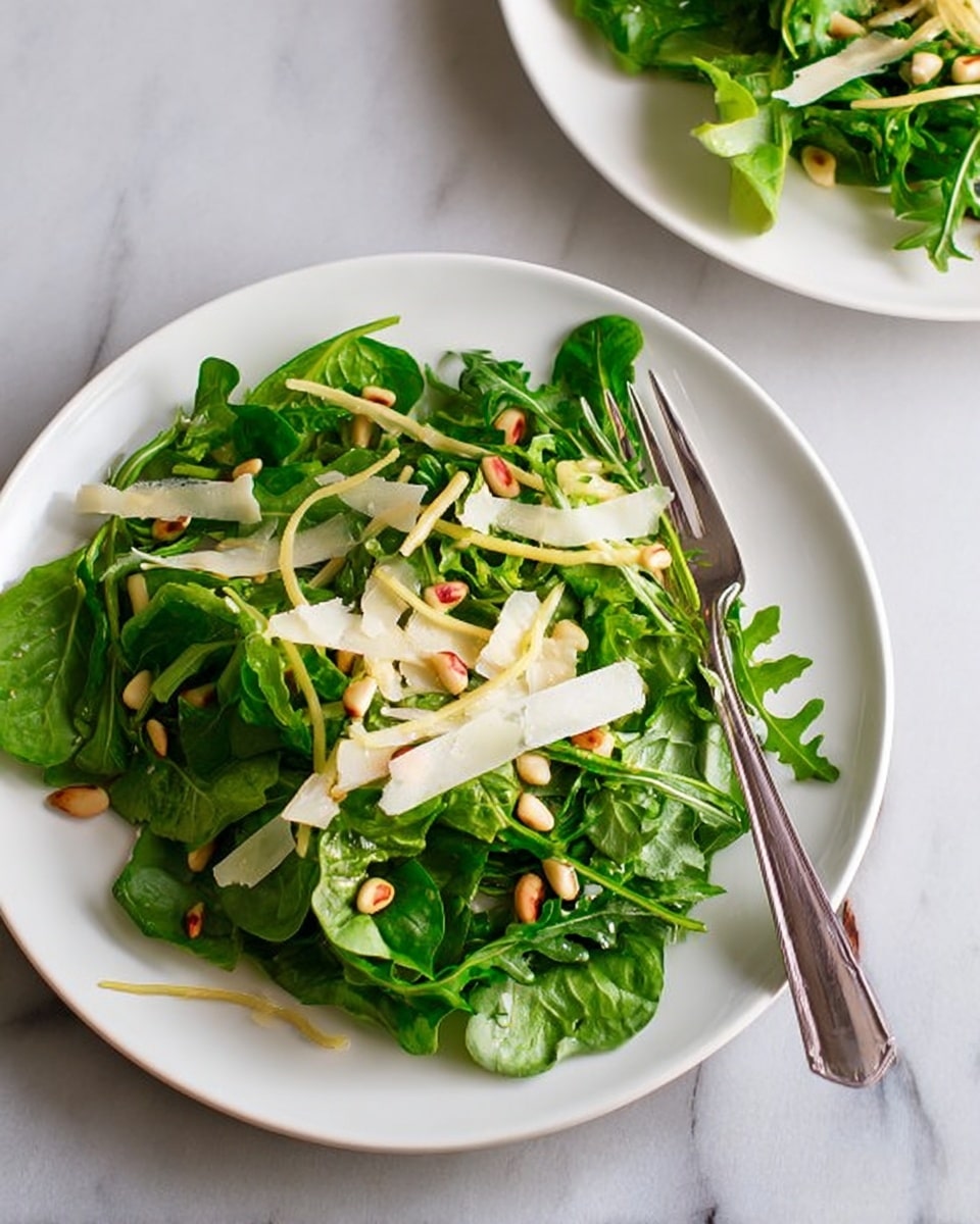 The image shows a white plate filled with a fresh green salad. The bottom layer is made of dark and bright green leafy vegetables, including arugula. On top of the greens, there are thin light yellow strips that look like shaved cheese or a vegetable. Scattered on top are small, light brown pine nuts that add texture. To the left side of the plate, next to the salad, is a silver fork resting on the white plate. The background is a white marbled texture surface. Photo taken with an iphone --ar 4:5 --v 7
