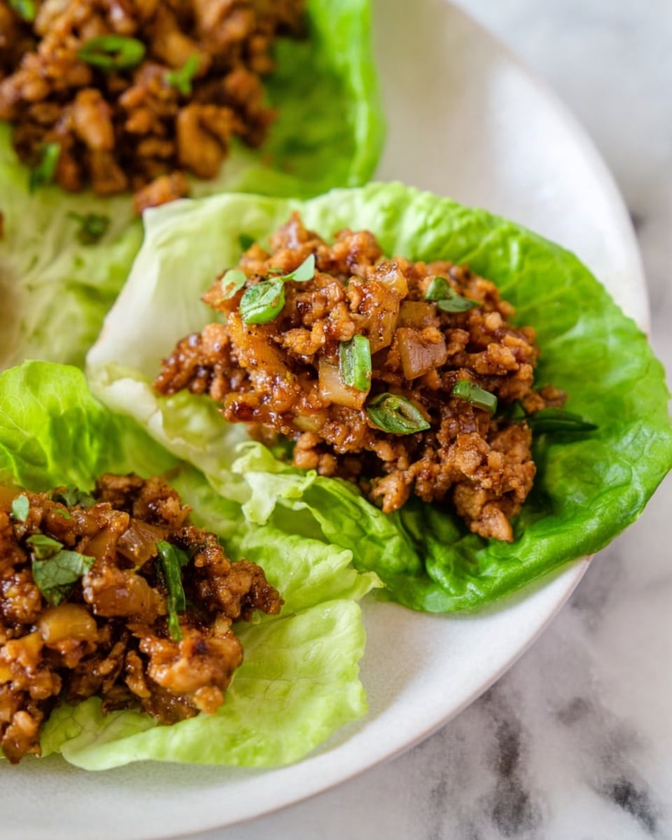 The image shows white plate with three lettuce wraps placed on a white marbled surface. Each wrap has two main layers: a bright green, fresh, and crinkly lettuce leaf forming the base, and on top, a textured mound of chopped cooked onions and finely chopped brown cooked meat, with a slight shiny glaze, garnished by a few small green herb pieces scattered on top. The colors stand out with the light green of the lettuce contrasting with the warm, deep brown of the meat and onions. Photo taken with an iphone --ar 4:5 --v 7