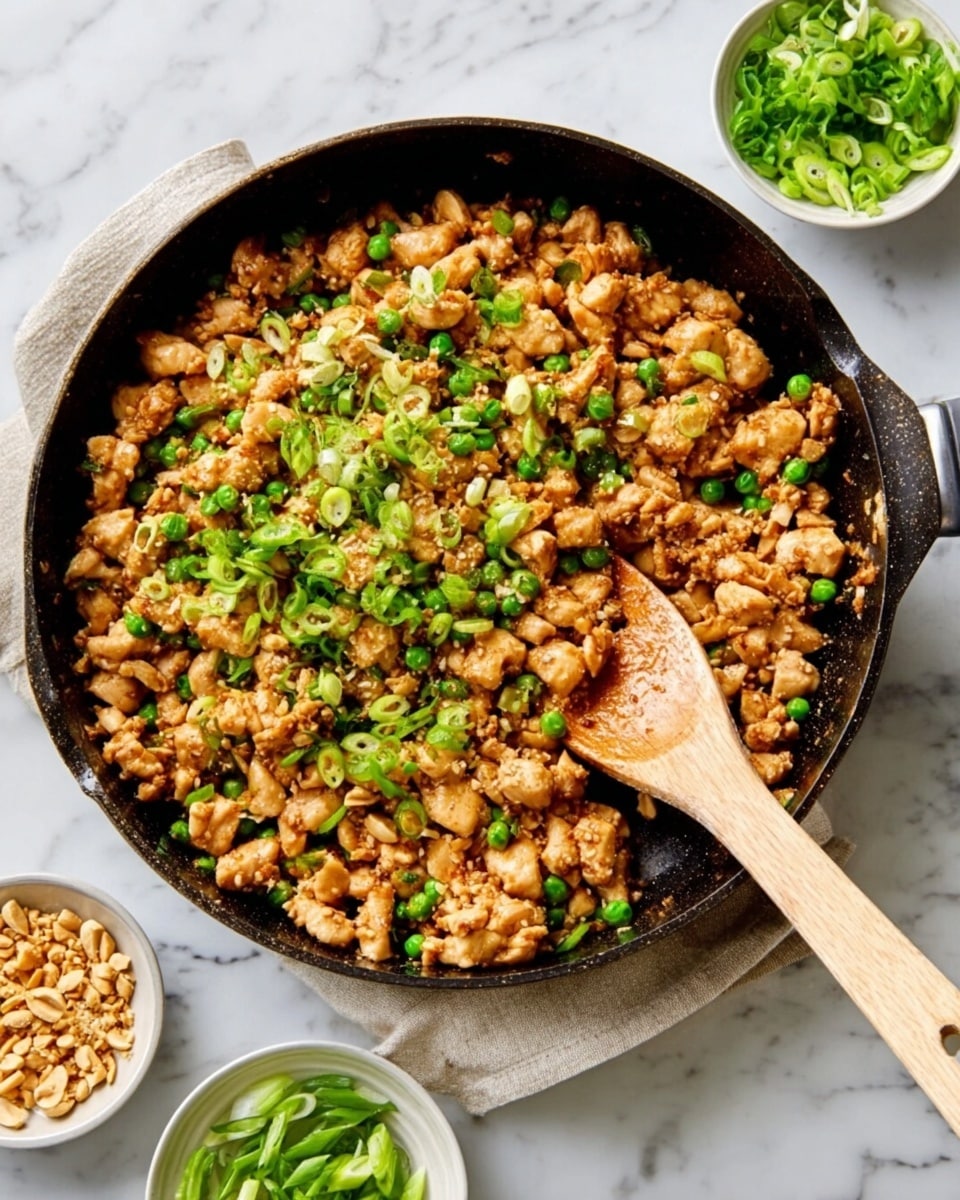A black pan filled with cooked small pieces of light brown chicken mixed with green peas and sprinkled with chopped green onions on top. A wooden spoon with a pale handle rests inside the pan, partly submerged in the chicken mixture. Around the pan on a white marbled surface, there are small white bowls each holding chopped green onions and crushed peanuts. Photo taken with an iphone --ar 4:5 --v 7