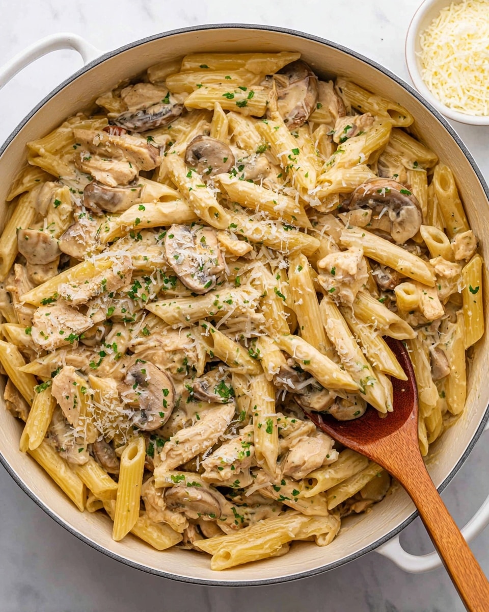 A large white pot filled with creamy penne pasta mixed with sliced mushrooms and shredded light tan chicken pieces. The pasta is coated evenly in a light beige creamy sauce with small bits of grated cheese on top. Sprinkled green herbs are scattered across the pasta for a fresh look. A wooden spoon is scooping some pasta from the right side, showing the thick, creamy texture. The pot is placed on a white marbled surface, with a small white bowl of grated cheese in the top right corner. photo taken with an iphone --ar 4:5 --v 7