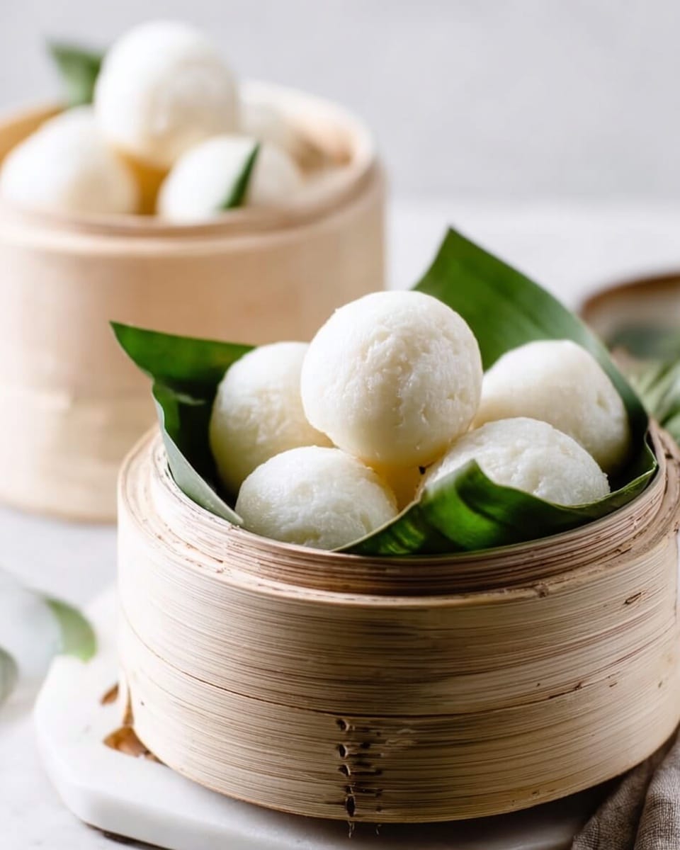 The image shows a round light bamboo steamer basket filled with soft white round dumplings arranged on green banana leaves inside. The dumplings have a smooth, slightly shiny texture and are stacked with some on top of others in a single layer inside the basket. The steamer sits on a white marbled surface with a soft light background and there is a second similar steamer with dumplings slightly blurred in the background. Photo taken with an iphone --ar 4:5 --v 7