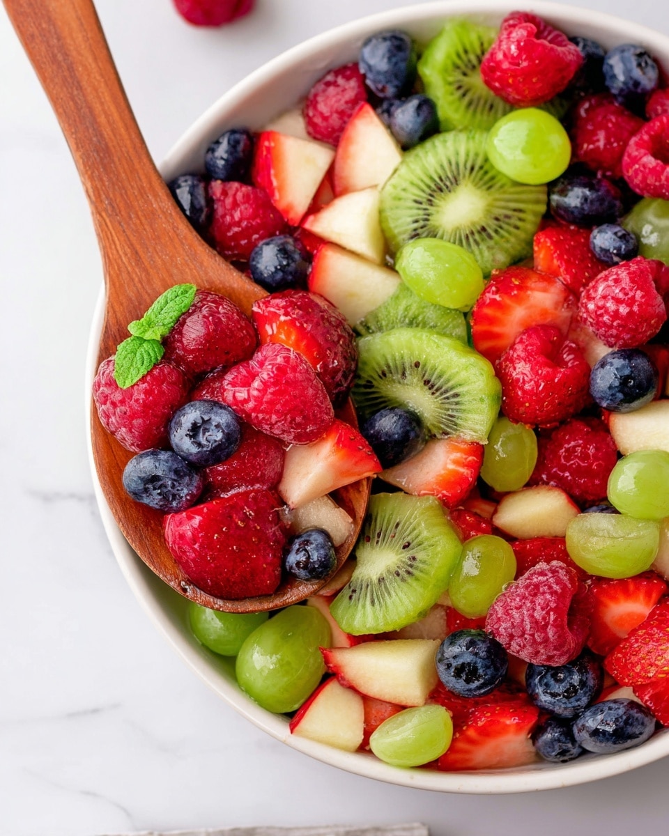 A white bowl filled with a colorful fruit salad showing many layers of fresh fruit. The base layer has green grape slices scattered around. Above them, there are many whole blueberries, red raspberries, and sliced strawberries with red and white tones. On top, pieces of green kiwi and small apple slices with red skin are mixed in. A wooden spoon rests on the bowl's edge, lifting some of the fruit. The fruit looks shiny and fresh, and a small green leaf sits among the berries for decoration, all placed on a white marbled surface. photo taken with an iphone --ar 4:5 --v 7