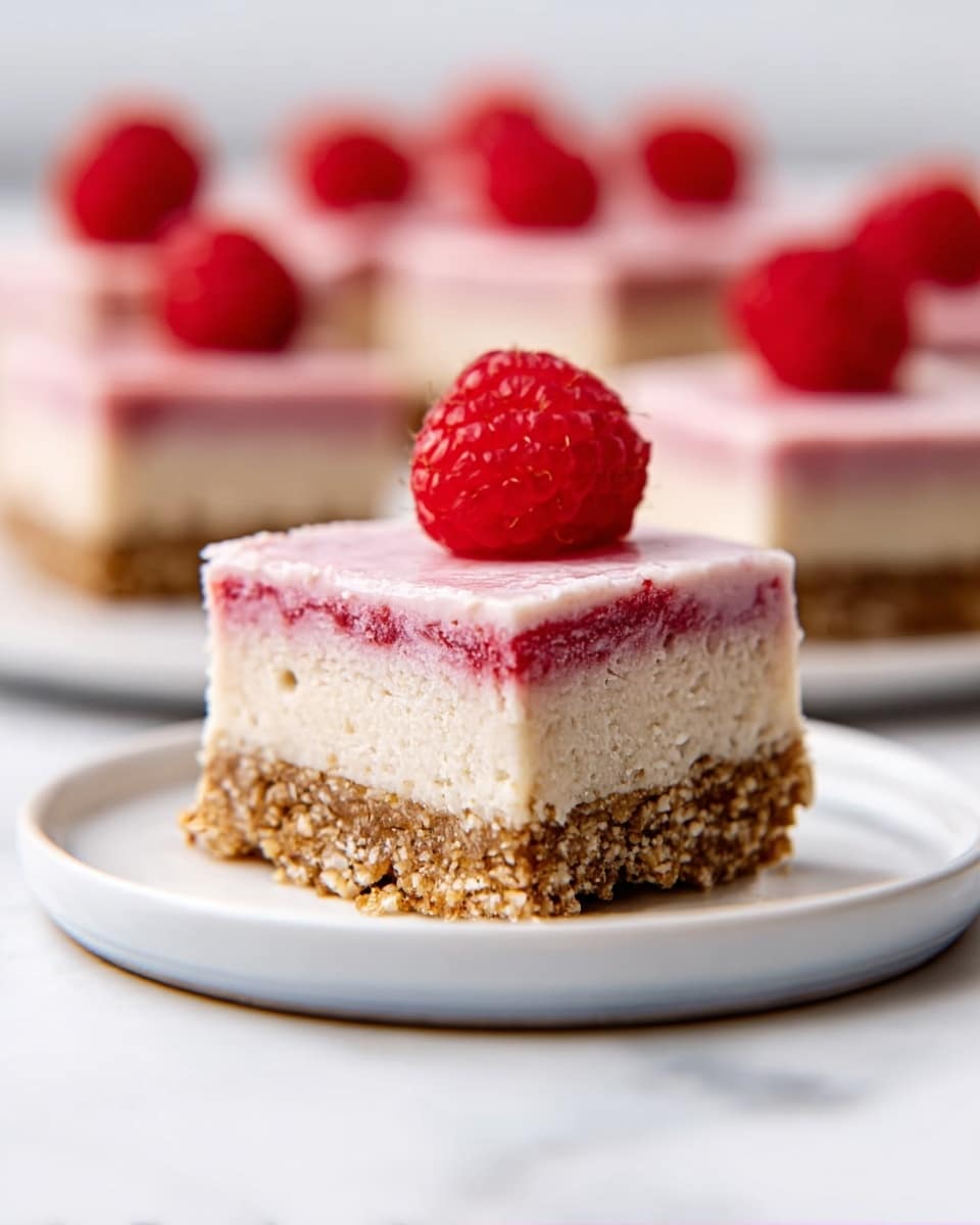 The image shows a small square cake slice on a white plate set on a white marbled surface. The cake has three clear layers: the bottom layer is a dense, crumbly brown base like crushed nuts or biscuits, the middle layer is creamy and light pink with a smooth texture, and the top layer is slightly lighter creamy white, also smooth. A fresh red raspberry sits on top of the cake, adding a pop of color. In the background, several similar cake slices are blurred, showing consistent layering and raspberries. The lighting is bright and natural, highlighting the textures and colors. Photo taken with an iphone --ar 4:5 --v 7