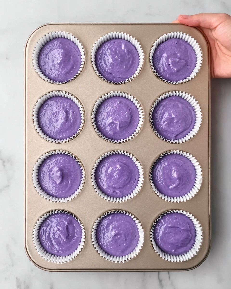 A baking tray with twelve white paper cupcake liners filled with smooth purple batter. Each liner is evenly filled, showing a shiny, wet surface with slight texture from the batter. The tray is placed on a white marbled countertop, and a woman's hand is holding the tray from the top left corner. The scene is bright and clean, focusing on the vibrant purple color of the batter and the neat layout of the liners. photo taken with an iphone --ar 4:5 --v 7