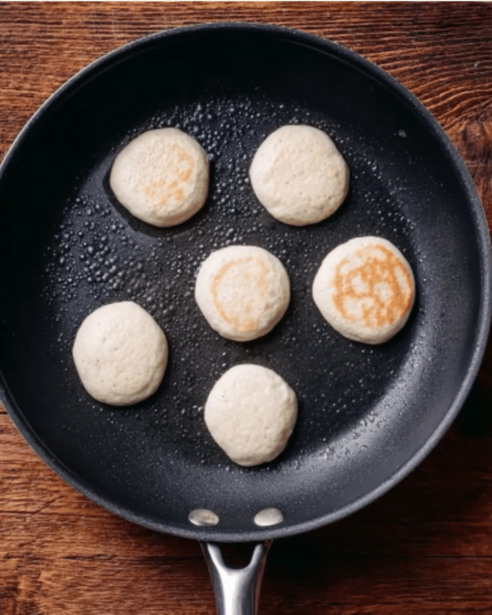 A black frying pan contains seven small, round pancakes cooking. Each pancake is pale cream in color, with a smooth texture and a few small bubbles on the surface. The pancakes are arranged spaced apart inside the pan. The pan is set on a wooden surface. photo taken with an iphone --ar 4:5 --v 7
