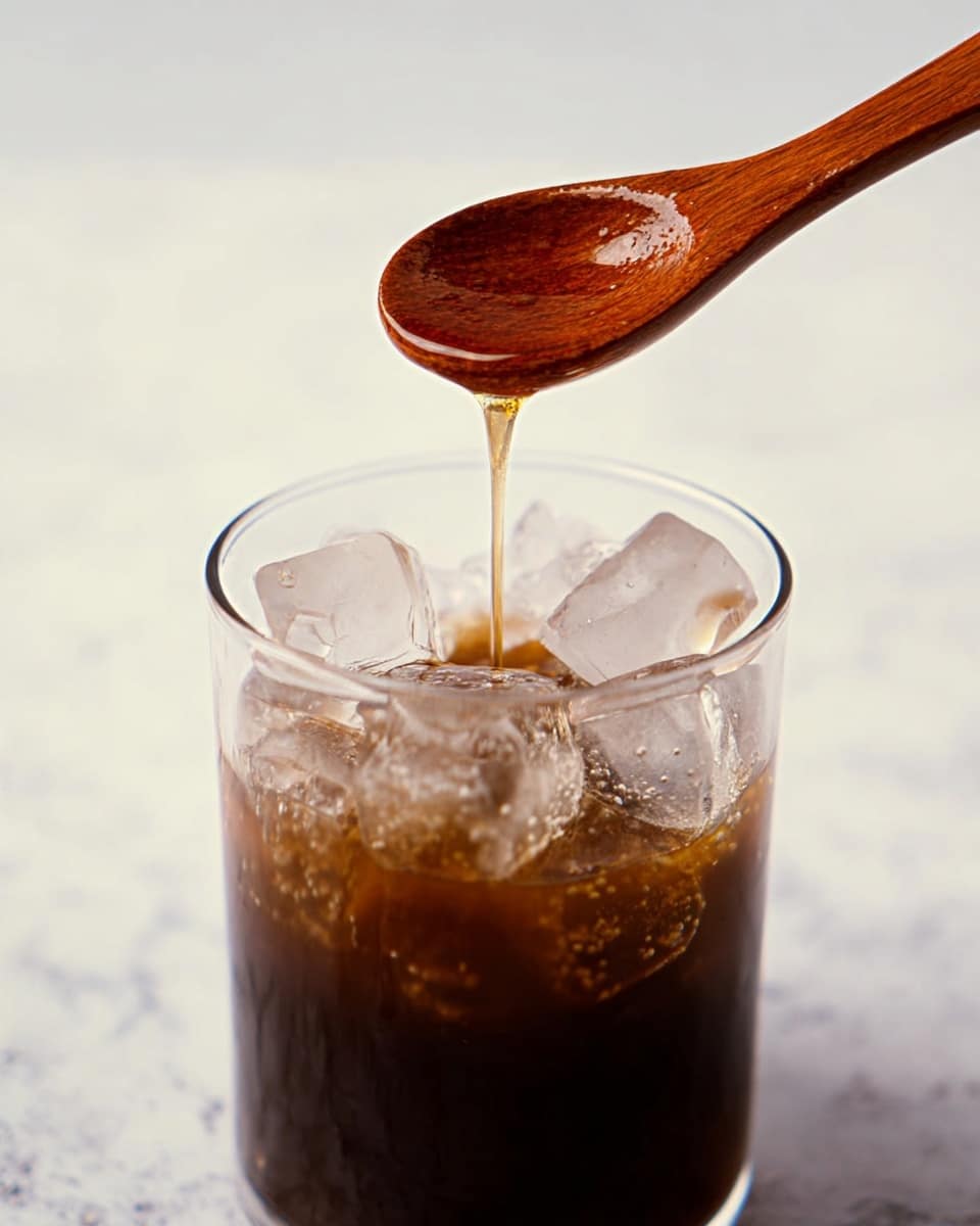 A clear glass filled halfway with dark brown liquid and topped with irregular shaped ice cubes that are mostly translucent with some white frost. Above the glass, a wooden spoon with a rich brown polished texture is held slightly tilted, dripping a thin stream of light honey-colored syrup into the glass. The background is a white marbled texture, giving a clean and bright look. photo taken with an iphone --ar 4:5 --v 7