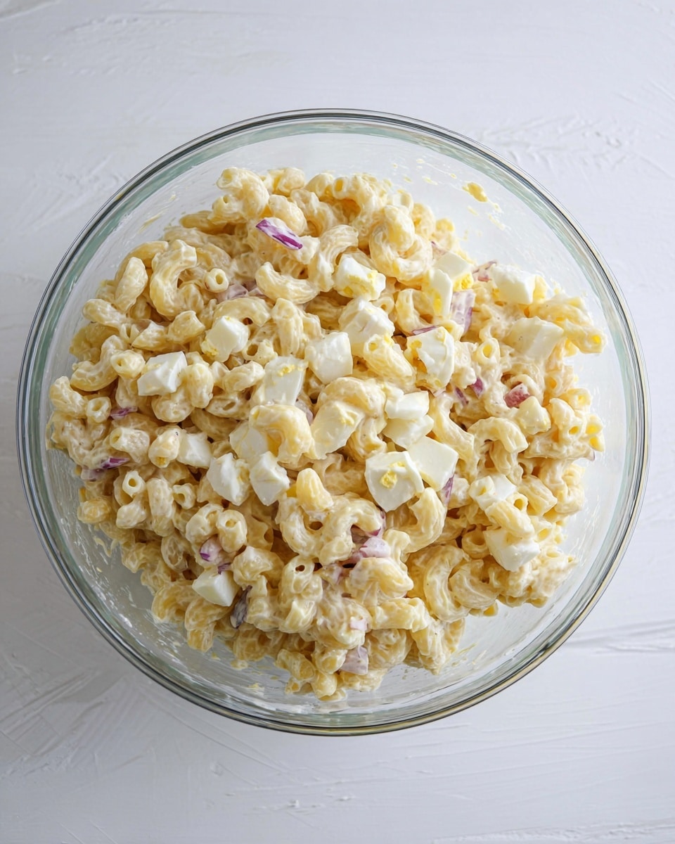 A large clear glass bowl filled with a creamy macaroni salad, showing about three main layers. The top layer has small pieces of white and pale yellow chopped boiled eggs mixed evenly with pale beige curved macaroni pasta coated in a light creamy dressing. Tiny bits of purple red onion are scattered throughout, adding slight color contrast among the smooth and soft texture of the pasta and eggs. The bowl sits on a white marbled surface, enhancing the pale tones of the salad. photo taken with an iphone --ar 4:5 --v 7