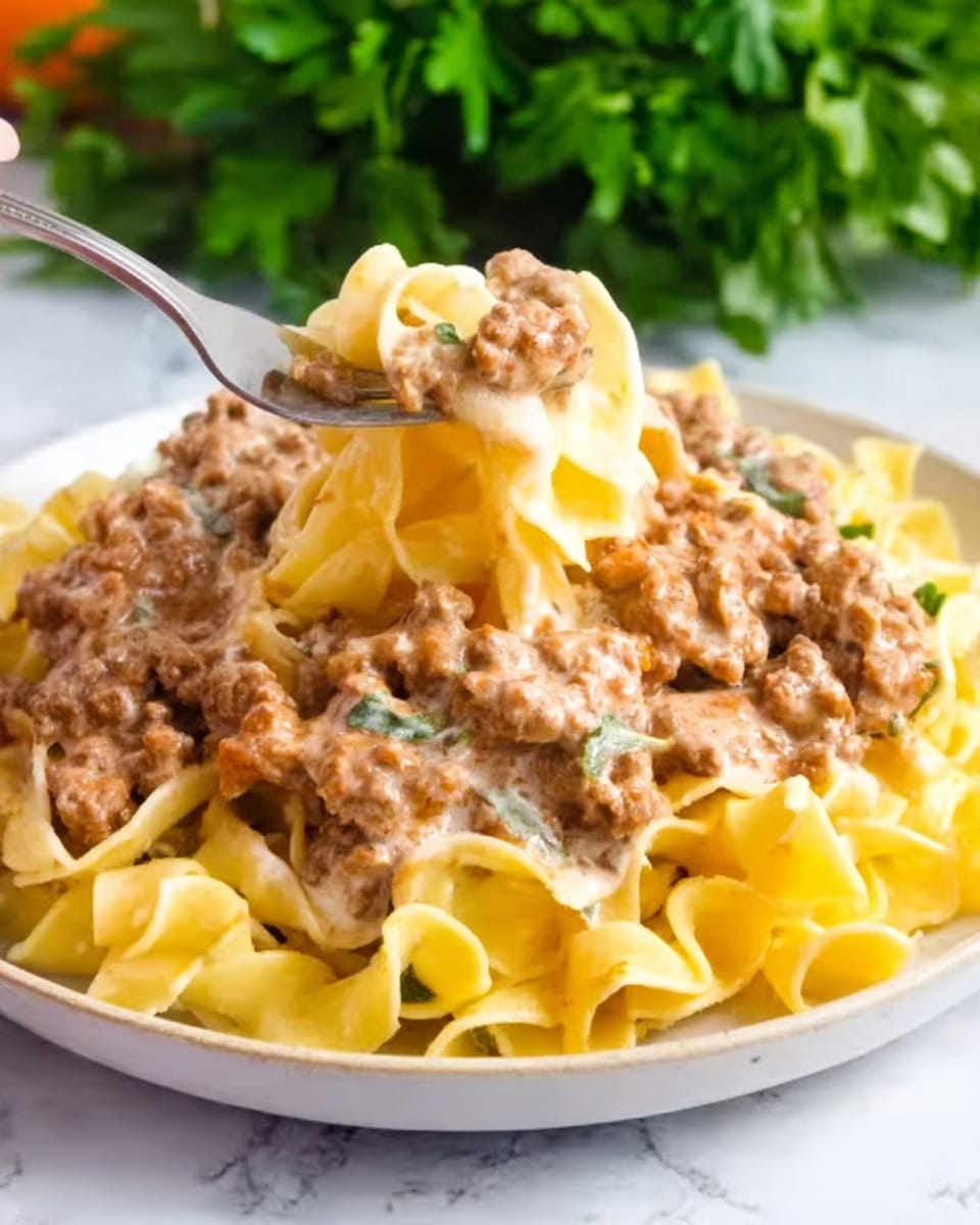 The image shows a white bowl filled with a creamy beef and mushroom stroganoff dish. At the bottom, there is a layer of soft buttered egg noodles with a light golden color and slightly curled edges. On top of the noodles, pieces of brown cooked beef strips and sliced mushrooms are spread evenly, all coated in a thick, rich brown sauce with specks of green herbs. The bowl is placed on a white marbled surface, with part of another white bowl containing sour cream visible in the background. photo taken with an iphone --ar 4:5 --v 7