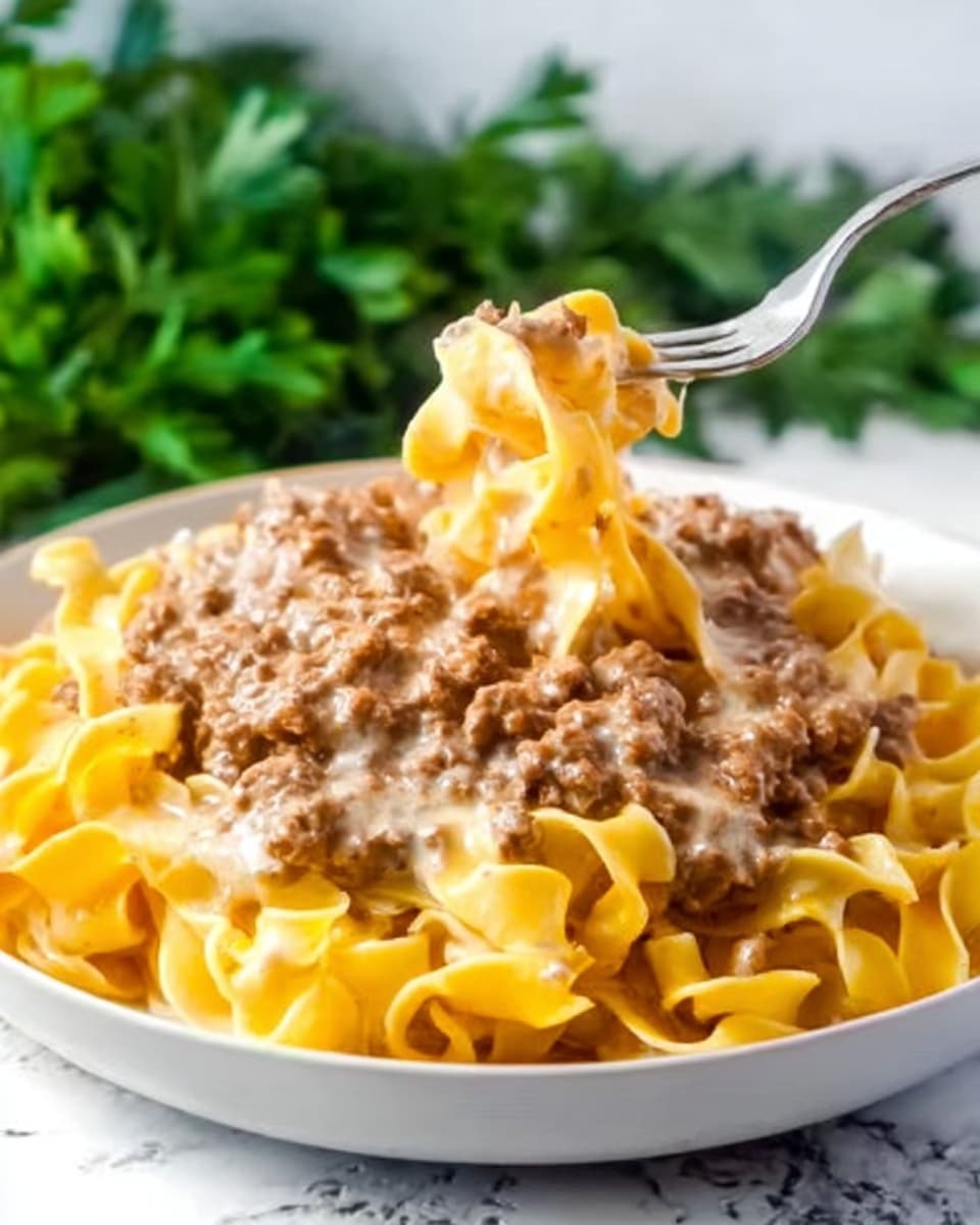 The dish shows a white plate filled with wide, yellow egg noodles forming the base layer, topped with a thick layer of creamy sauce mixed with cooked ground meat that looks light brown and soft in texture. A woman's hand with a silver fork is lifting some of the pasta and meat sauce, revealing the smooth and rich sauce coating each noodle. The background is a white marbled texture with blurred green leafy herbs in the back, giving a fresh contrast to the warm tones of the dish. photo taken with an iphone --ar 4:5 --v 7