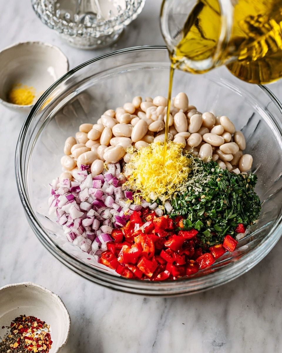 A clear glass bowl placed on a white marbled surface is filled with layers of ingredients for a dish: a large base of white beans surrounds finely chopped raw red onions on the left, bright red diced peppers on the bottom right, and a fresh pile of chopped green herbs in the center. On top of the herbs sits a small mound of yellow lemon zest and a sprinkle of coarse salt, black pepper, and red chili flakes. A woman's hand is pouring golden olive oil from a clear glass container into the bowl from the right side. In the background, there are small white dishes holding extra seasonings and a clear glass juicer with a lemon half resting in it. photo taken with an iphone --ar 4:5 --v 7