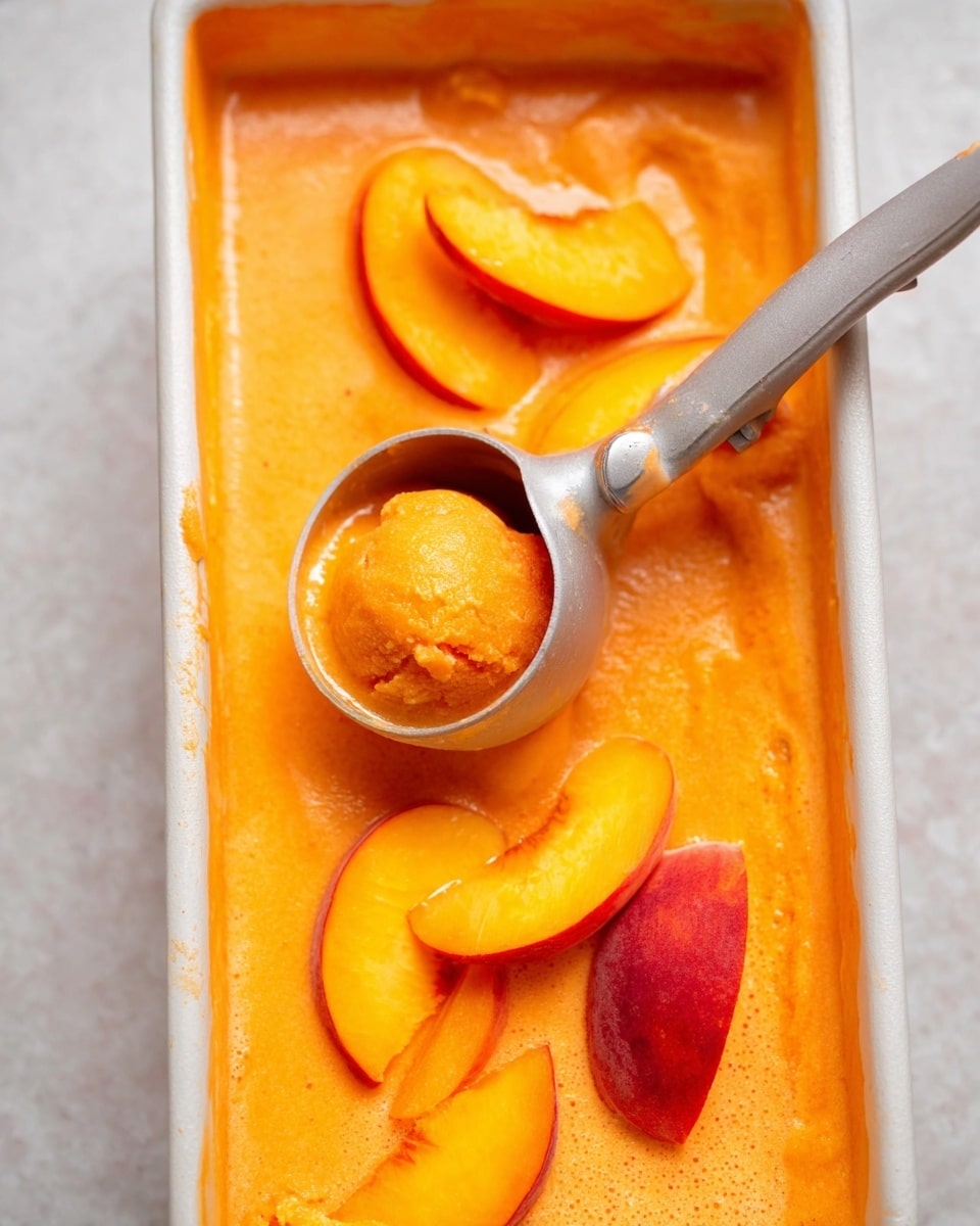 The image shows a long rectangular container filled with bright orange creamy mixture. Inside the mixture, a silver ice cream scoop is lifting a small ball of the orange frozen dessert. Around the scoop, there are several slices of fresh peach with yellow-orange skin and soft texture resting on the mixture. The background surface is a white marbled texture. photo taken with an iphone --ar 4:5 --v 7