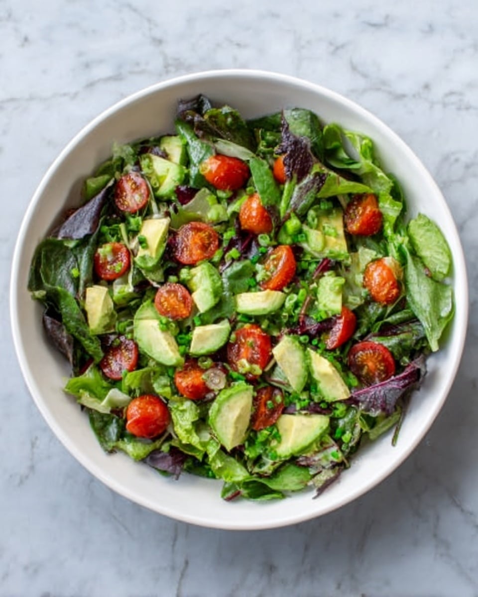A white bowl filled with a fresh green salad sits on a white marbled surface. The salad has multiple layers starting with dark and light green leafy vegetables like spinach and lettuce. Scattered across the greens are bright red cherry tomatoes, whole and sliced. There are also pieces of light green avocado and chopped crisp green onions mixed throughout. The colors are vibrant, with the reds standing out against the greens and the textures ranging from smooth avocado to juicy tomatoes and leafy greens. Photo taken with an iphone --ar 4:5 --v 7