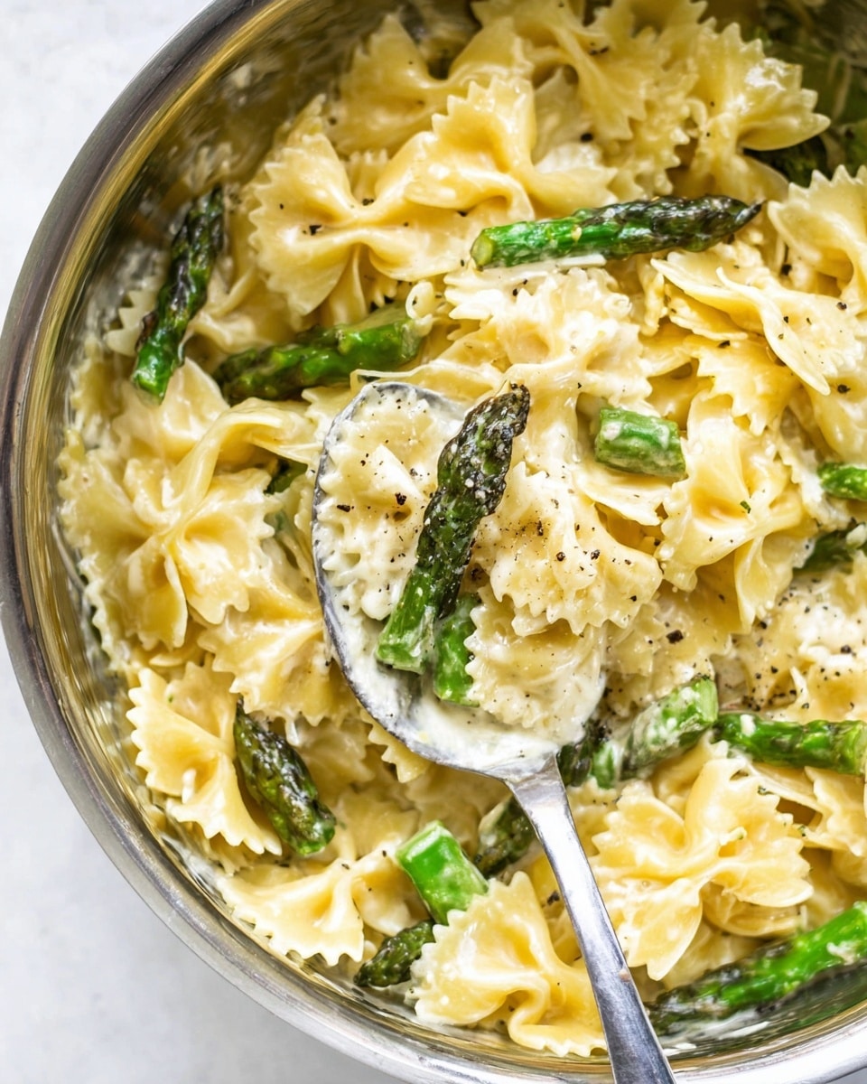 A close-up view of a metal bowl filled with creamy farfalle pasta layered with bright green asparagus pieces. The farfalle pasta is light yellow and covered with a smooth cream sauce, sprinkled lightly with black pepper and grated cheese. A large silver spoon rests inside the bowl, holding some pasta and sauce, showing the thick texture of the cream. The dish sits on a white marbled textured surface. photo taken with an iphone --ar 4:5 --v 7