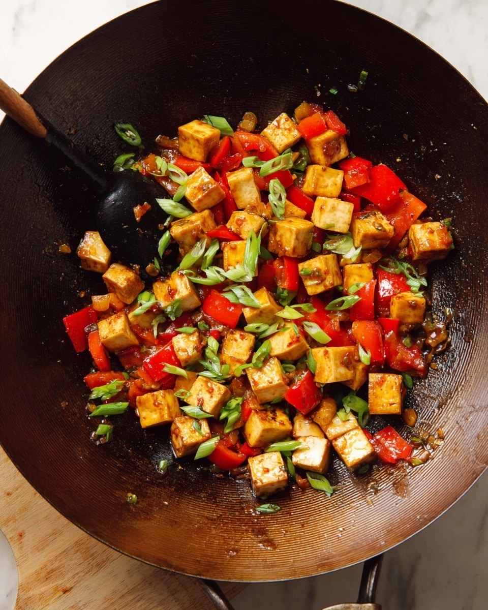 The image shows a large dark brown wok filled with small cooked golden brown tofu cubes mixed with bright red bell pepper pieces and scattered slices of green onions on top. The tofu and bell peppers have a shiny, slightly saucy texture, and there is a black spoon stirring the mix inside the wok. The wok rests on a white marbled surface with light wooden tones visible underneath. Photo taken with an iphone --ar 4:5 --v 7