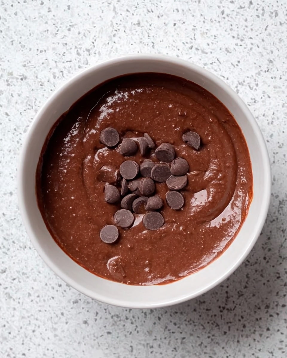 The image shows a white bowl filled with smooth, thick chocolate batter. There are small, round chocolate chips scattered unevenly on the surface of the batter. The bowl is placed on a white marbled textured surface, making the dark brown color of the chocolate stand out clearly. The scene is simple with a focus on the rich, glossy texture of the chocolate mixture. photo taken with an iphone --ar 4:5 --v 7