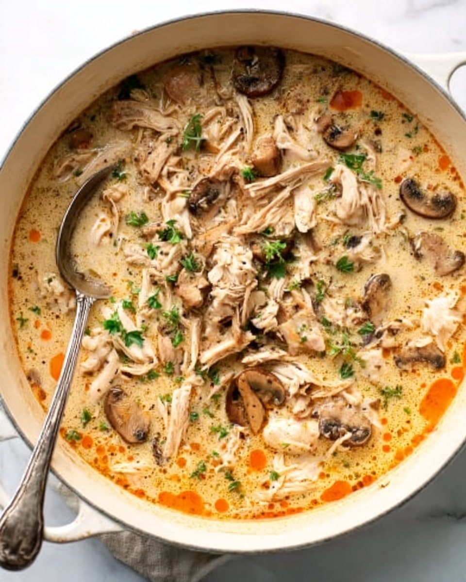 A white pot filled with creamy soup that has visible sliced mushrooms and shredded pieces of chicken spread evenly on top. The soup base is light beige with orange oil spots floating around the edges. Small green herbs are sprinkled throughout, adding a touch of color. A metal spoon rests inside the pot on the left side. The background shows a white marbled surface. Photo taken with an iphone --ar 4:5 --v 7