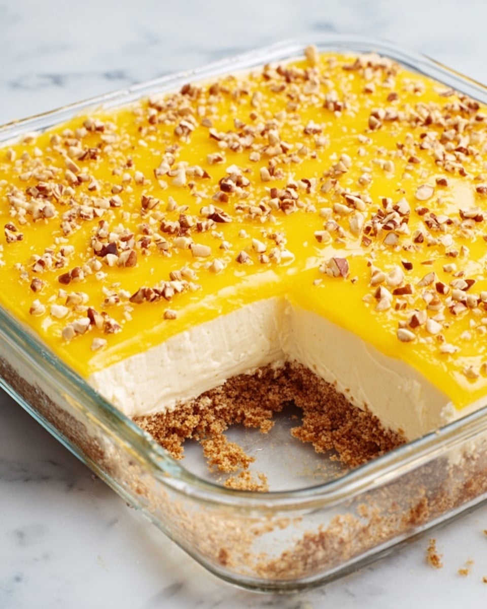 A clear glass square dish filled with a three-layer dessert: the bottom layer is crumbly and brown, the middle layer is smooth and creamy white, and the top layer is bright yellow with a shiny texture, sprinkled with small chopped nuts spread evenly across. A portion of the dessert has been removed, showing the layers clearly. The dish is placed on a white marbled surface. Photo taken with an iphone --ar 4:5 --v 7