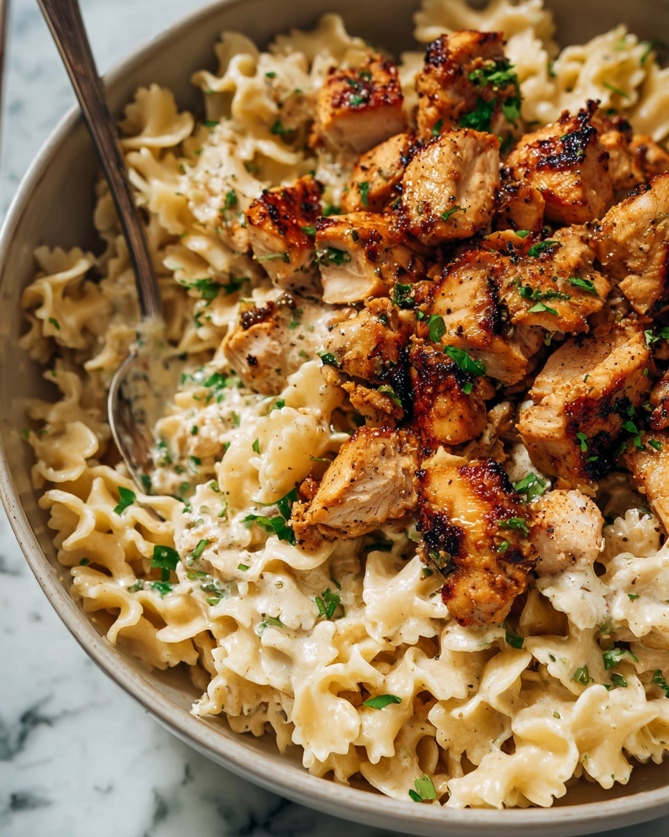 The image shows a close-up of a creamy pasta dish with two main layers. The bottom layer is light cream-colored pasta with ruffled edges, soft in texture and coated in a smooth sauce. On top are pieces of golden-brown grilled chicken with a slightly crispy surface, broken into bite-sized chunks. Small green herb pieces are sprinkled over the dish, adding a fresh touch. The dish is served in a white bowl on a white marbled surface, and there's a silver spoon partially visible. Photo taken with an iphone --ar 4:5 --v 7