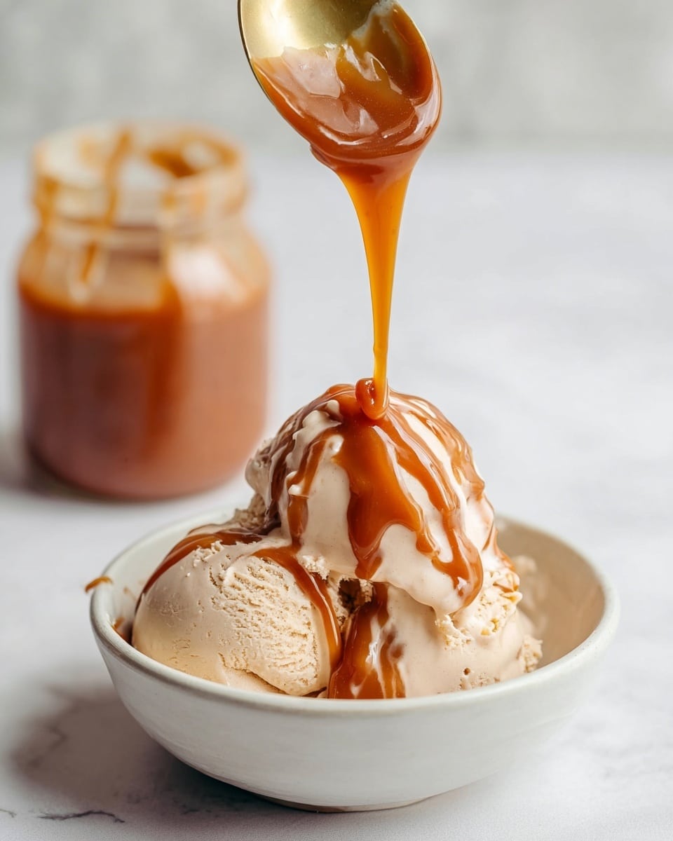 The image shows three scoops of light beige ice cream in a white bowl on a white marbled surface. A thick, shiny caramel sauce with a rich brown color is being poured over the top scoop from a golden spoon held by a woman’s hand, creating glossy streaks and drips that flow down the ice cream, adding a smooth and sticky texture. In the background, a glass jar filled with more caramel sauce is softly blurred, complementing the scene. photo taken with an iphone --ar 4:5 --v 7