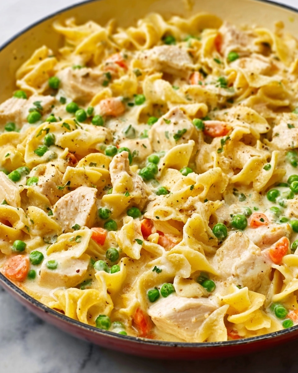 This image shows a close-up of a creamy pasta dish with three layers visible. The base layer consists of wide, twisted egg noodles that are golden yellow with a soft texture. On top of the noodles are chunks of light tan cooked chicken mixed with small bright green peas and small pieces of orange carrots, scattered evenly throughout. The entire dish is covered with a thick, creamy white sauce that glistens and fills in the gaps between the noodles and vegetables. The dish is presented in a white skillet, sitting on a white marbled surface. photo taken with an iphone --ar 4:5 --v 7