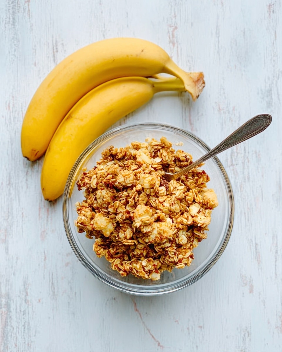 A clear glass bowl filled with golden brown granola sits on a white marbled surface. The granola looks crunchy with clusters of oats and small pieces mixed together. Next to the bowl, there are two bright yellow bananas resting closely on the surface. A silver spoon is placed inside the bowl, angled to the right side with its handle visible. The overall light is soft and natural, showing the textures clearly. Photo taken with an iphone --ar 4:5 --v 7