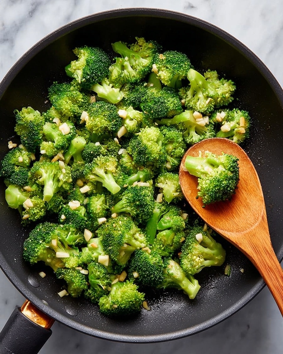 A black frying pan filled with bright green broccoli pieces that look fresh and slightly cooked, scattered with small white garlic chunks. A wooden spoon is inside the pan, holding some broccoli, resting near the middle right side. The pan sits on a white marbled surface. photo taken with an iphone --ar 4:5 --v 7