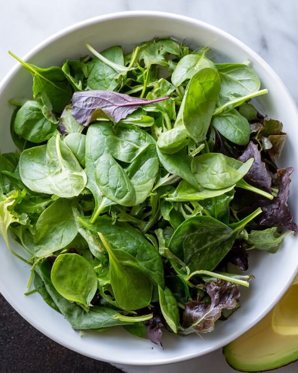 A white bowl filled with two layers of fresh salad greens, the bottom layer consisting of slightly darker leaves with a mix of green and purple shades, and the top layer made up of lighter bright green spinach leaves with smooth texture and curvy edges, all arranged loosely. The bowl sits on a white marbled surface, and part of a sliced avocado is visible at the right edge of the image. photo taken with an iphone --ar 4:5 --v 7