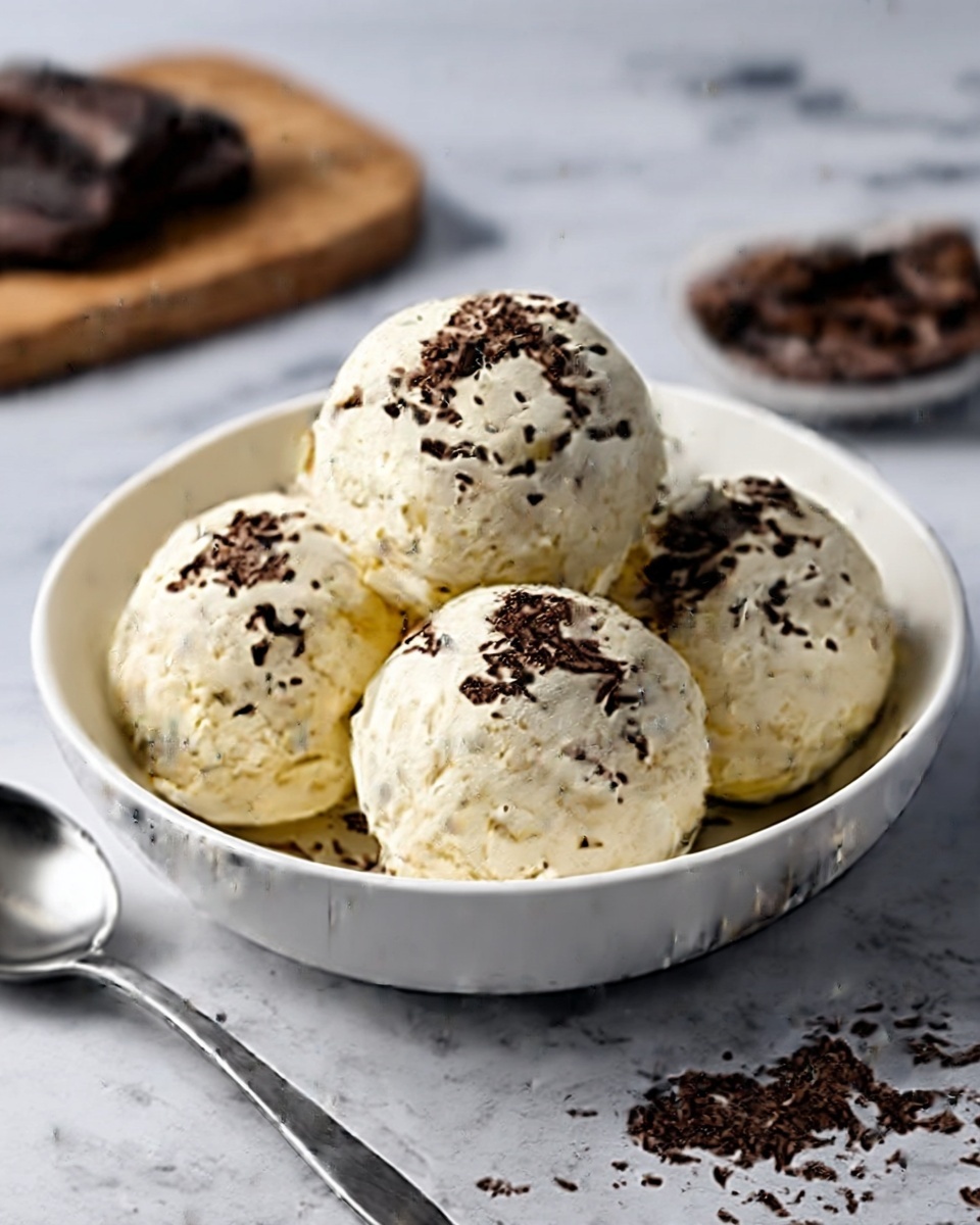 In a white bowl, there are four scoops of creamy vanilla ice cream piled closely together. Each scoop is smooth and slightly shiny, topped with small pieces of dark chocolate shavings scattered evenly over the ice cream. The bowl sits on a white marbled surface, with a silver spoon placed next to it on the left and a chopping board with more chocolate shavings blurred in the background. Photo taken with an iphone --ar 4:5 --v 7