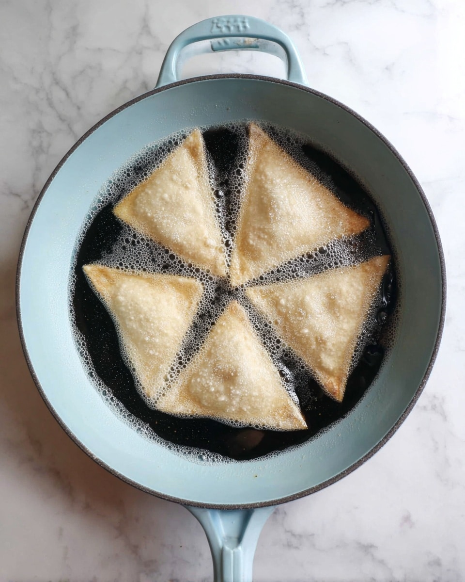 A light blue skillet filled with hot oil shows five light beige triangular pockets frying inside, each floating with a slightly puffed texture and some bubbles around the edges. The skillet sits on a white marbled surface, visible from above. Photo taken with an iphone --ar 4:5 --v 7