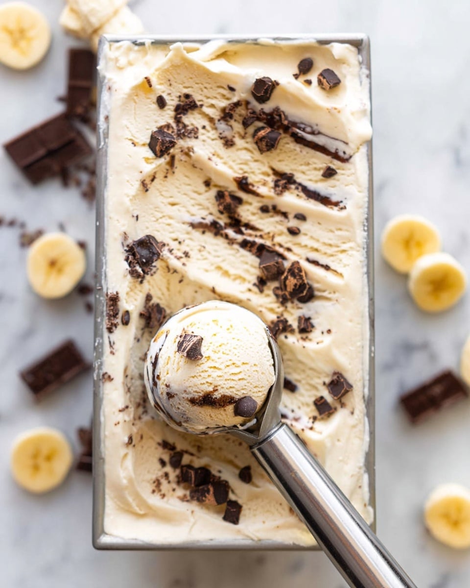 A close-up of a rectangular container filled with creamy beige ice cream mixed with scattered dark chocolate chunks throughout. The top layer is smooth with random swirls and small chocolate pieces spread on the surface. A metal ice cream scoop, held by a woman's hand, is embedded in the ice cream, taking out a round scoop that shows soft texture and studded chocolate bits inside. The container sits on a white marbled surface, and some banana slices and chocolate chunks are placed loosely around it. photo taken with an iphone --ar 4:5 --v 7