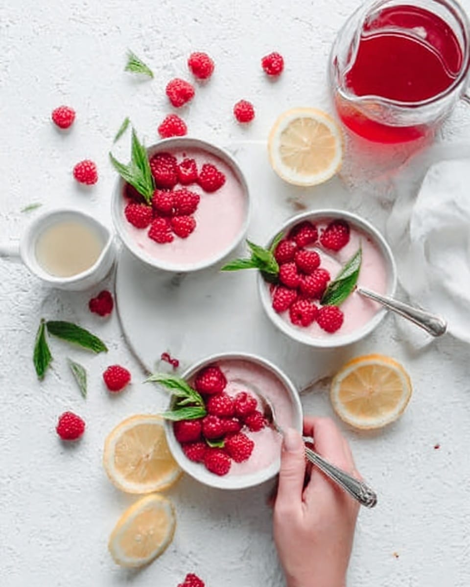 The image shows a white marbled surface with three white bowls filled with a dessert. Each bowl has one main layer of pale pink creamy texture topped with fresh bright red raspberries and a few green leaves for decoration. Next to the bowls, there is a small white pitcher filled with red sauce and a small white bowl with a light cream sauce. A woman's hand holds one of the bowls with a silver spoon inside. There are scattered lemon wedges and raspberries around the bowls. The scene is bright and fresh looking. Photo taken with an iphone --ar 4:5 --v 7